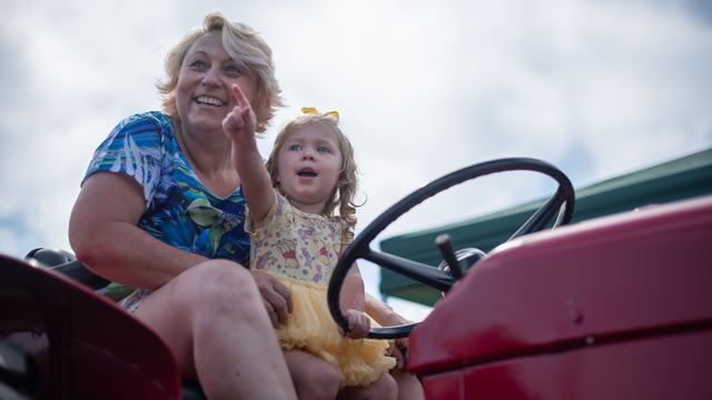 A woman and a young girl sit on a red tractor; the woman smiles and points toward the distance while the toddler looks on with an excited expression.