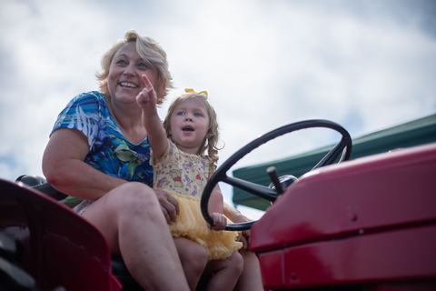 A woman and a young girl sit on a red tractor; the woman smiles and points toward the distance while the toddler looks on with an excited expression.