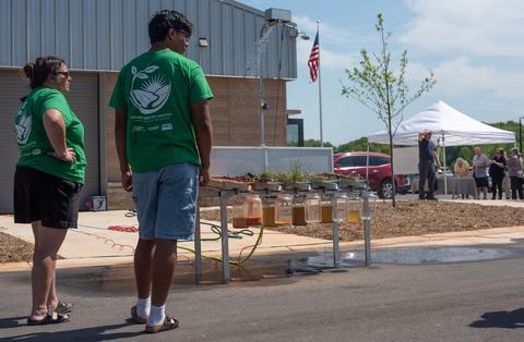 Two people in green Common Ground Festival shirts observe a soil erosion demonstration where water sprays over different soil samples into hanging glass jars.