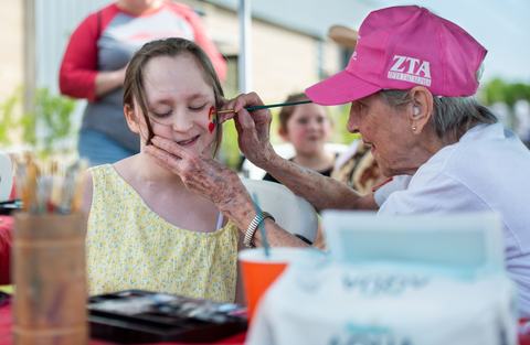 An elderly woman wearing a pink Zeta Tau Alpha hat paints a small red heart on the cheek of a smiling young girl in a yellow dress at an outdoor festival.