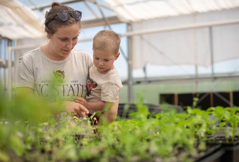 A woman wearing an NC State Wolfpack shirt holds a young boy as they look closely at green seedlings growing in a greenhouse.