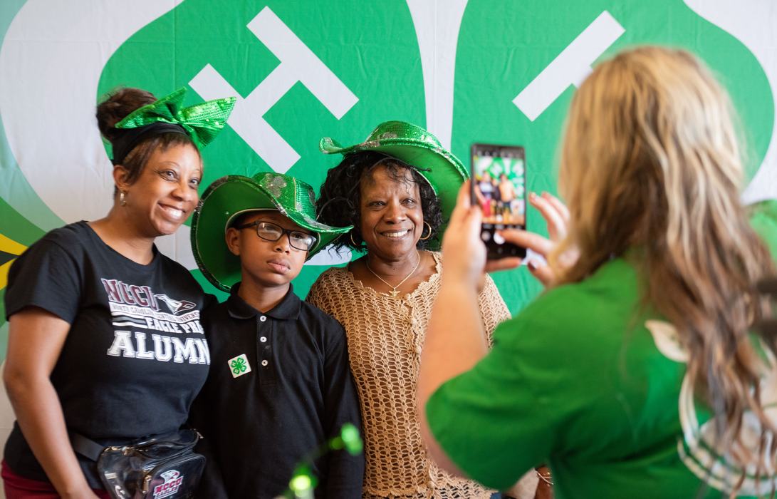 Three people—a woman in a 4-H alumni shirt, a young boy, and an older woman—pose for a photo in front of a large green 4-H clover backdrop. They are wearing festive green hats and accessories while a person in the foreground captures the moment on a smart