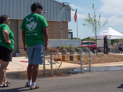 Two people in green Common Ground Festival shirts observe a soil erosion demonstration where water sprays over different soil samples into hanging glass jars.