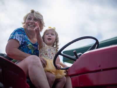 A woman and a young girl sit on a red tractor; the woman smiles and points toward the distance while the toddler looks on with an excited expression.