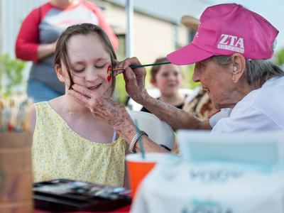 An elderly woman wearing a pink Zeta Tau Alpha hat paints a small red heart on the cheek of a smiling young girl in a yellow dress at an outdoor festival.