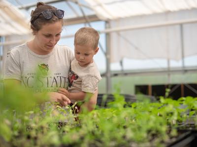 A woman wearing an NC State Wolfpack shirt holds a young boy as they look closely at green seedlings growing in a greenhouse.