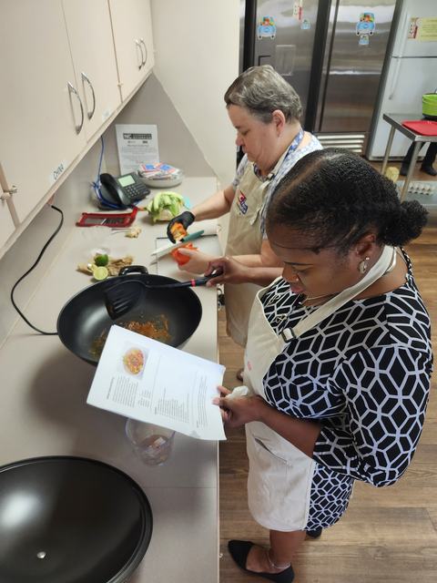Two adults stand at a counter, one is redaing the recipe while the other preps veggies to go in the electric stir fry skillet.