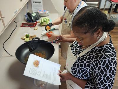 Two adults stand at a counter, one is redaing the recipe while the other preps veggies to go in the electric stir fry skillet.