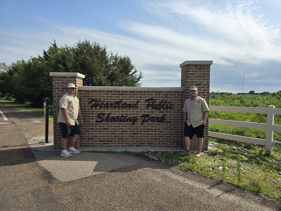 Two young men in matching tan shirts, dark shorts, and hats stand on either side of a brick entrance sign that reads "Heartland Public Shooting Park."