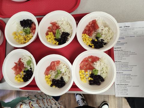 Several bowls full of grains, beans, corn, and tomatoes are lined up on a tray next to the recipe.