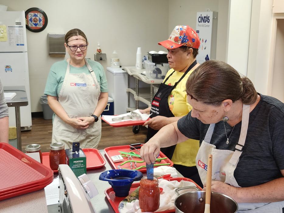 A Master Food Volunteer assists two participants with adding tomato sauce to jars during a tomato canning workshop.