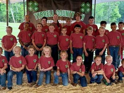A large group of children and teens wearing matching red T-shirts and blue jeans pose together in rows under a tent.  Behind them is a banner reading “Tyrrell County 4-H,” with green clover symbols, and a grassy outdoor area visible in the background.