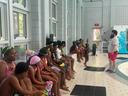 A group of children wearing swim caps and goggles sits along the edge of an indoor pool area while a instructor with a beard addresses them.