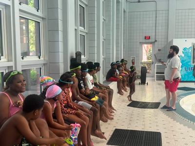 A group of children wearing swim caps and goggles sits along the edge of an indoor pool area while a instructor with a beard addresses them.