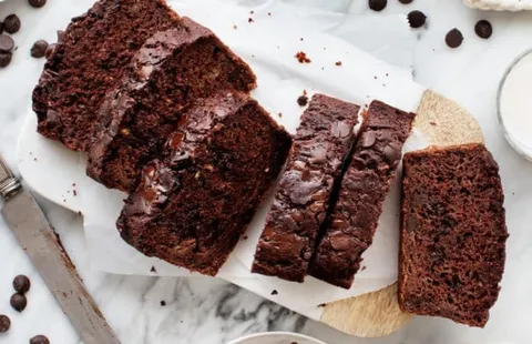 Chocolate zucchini bread sliced on a wooden cutting board, showing a moist texture with melted chocolate chips throughout. The loaf is surrounded by scattered chocolate chips and baking ingredients on a light countertop.