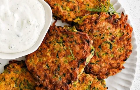 Golden-brown zucchini fritters with visible shredded zucchini and herbs, served on a white plate alongside a bowl of creamy dipping sauce and garnished with fresh dill.