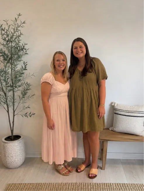 Two Halifax County 4-H agents standing together indoors, smiling for a photo. One is wearing a light pink dress, and the other is wearing an olive green dress.
