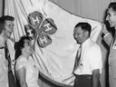 Black-and-white historical photograph of five 4-H club members standing together and smiling in front of a large 4-H flag during North Carolina State 4-H Club Week.