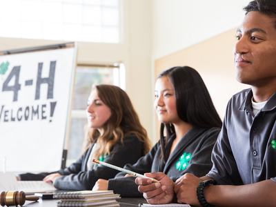 Three 4-H youth sit at a table during a meeting, wearing shirts with the 4-H clover emblem. A “4-H Welcome!” sign is visible in the background, along with small 4-H and American flags on the table.