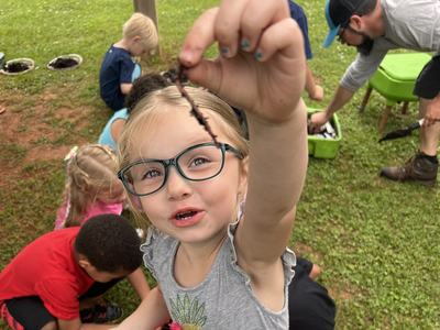 little girl holding up composting worm with other kids in the background exploring compost