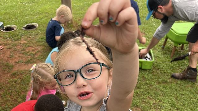 little girl holding up composting worm with other kids in the background exploring compost