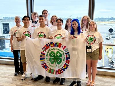 4-H youth group holding a States' International Exchange flag at an airport.