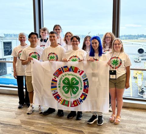 4-H youth group holding a States' International Exchange flag at an airport.