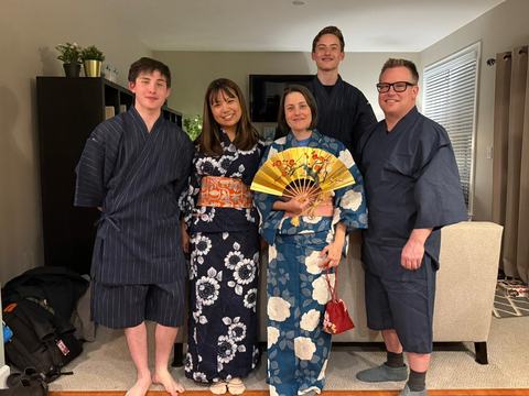 A host family poses with their exchange student from Japan. The host mom and student are wearing yukatas, and the host dad and brothers are wearing jimbei, traditional Japanese summer clothing. The host mother is holding a gold fan with a floral design.