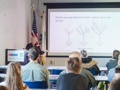 An EMG volunteer of Durham County presents on pruning in front of a presentation screen that shows the gardening tool in front of a classroom of volunteers who are focused on learning.