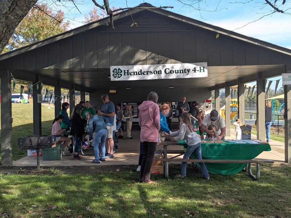 A crowd gathers under a park pavilion signed "Henderson County 4-H" for outdoor activities.