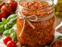A glass jar filled with roasted tomato pesto sits on a wooden surface, tied with rustic twine. Fresh tomatoes, basil leaves, garlic, and a bottle of olive oil surround the jar, highlighting ingredients used in the pesto.