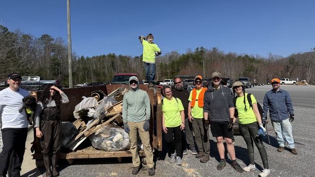 Lake James Clean Up 2026 volunteers standing in front of a dumpster full of trash including boards and tires. One of the volunteers in holding a dog and a child in standing on top of the dumpster.