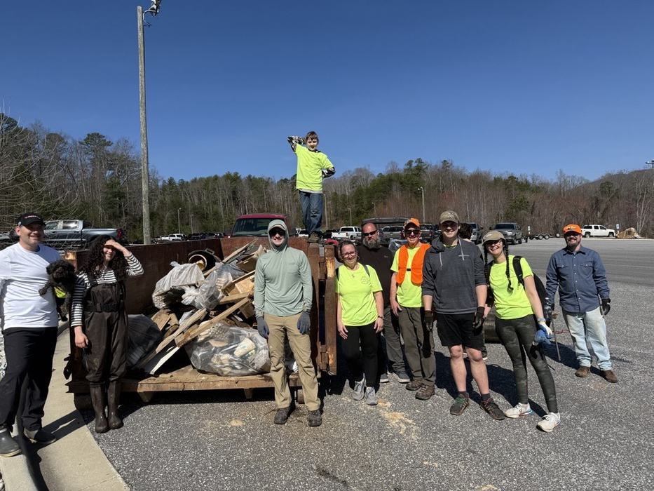 Lake James Clean Up 2026 volunteers standing in front of a dumpster full of trash including boards and tires. One of the volunteers in holding a dog and a child in standing on top of the dumpster.