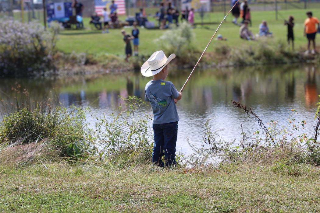 A young child in a cowboy hat stands by the edge of a pond holding a fishing rod.