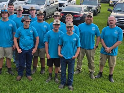 A group of young men and coaches wearing matching bright blue shirts and hats pose together outdoors on a grassy field with parked vehicles behind them.