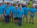 A group of young men and coaches wearing matching bright blue shirts and hats pose together outdoors on a grassy field with parked vehicles behind them.