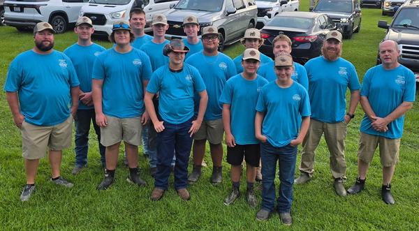 A group of young men and coaches wearing matching bright blue shirts and hats pose together outdoors on a grassy field with parked vehicles behind them.