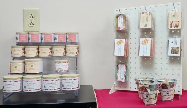 Handmade scented candles and wax melts arranged on tiered clear display stands and a white pegboard.