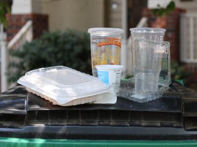 Assortment of clean, recyclable plastic food containers and cups on top of a bin. Shrub and front porch of home in the foreground.