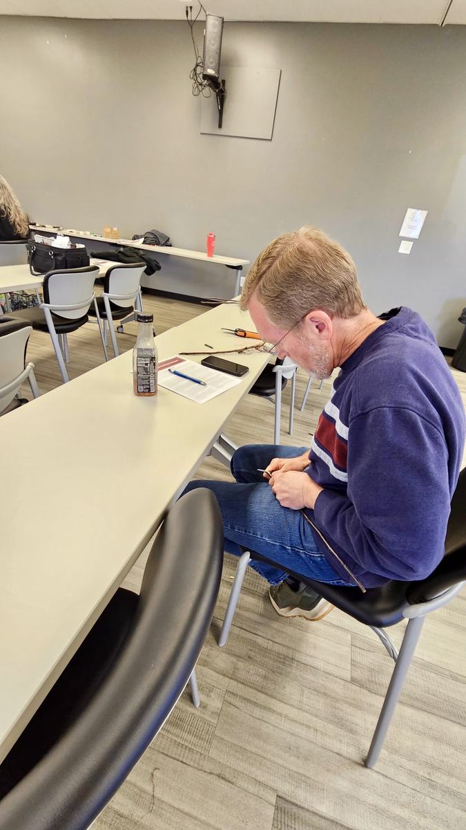 Participant seated at a classroom table carefully trimming apple scion wood with a grafting knife during a fruit tree grafting workshop at the Clay County Center.