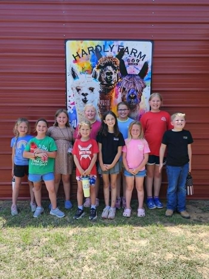 Ten children pose in front of a Hardly Farm sign with alpaca artwork during a field trip to the farm in Castalia, NC.