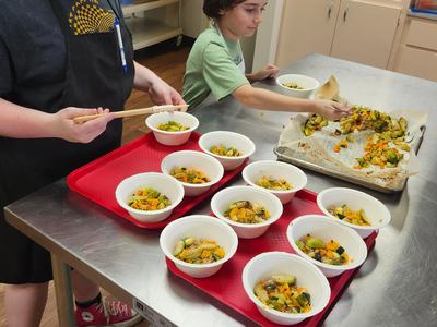 Two youth add roasted vegetables to 12 bowls filled with rice.