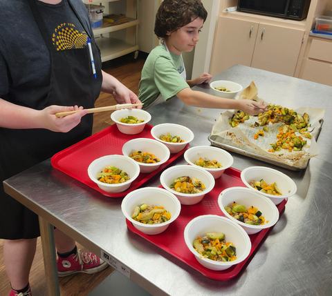 Two youth add roasted vegetables to 12 bowls filled with rice.