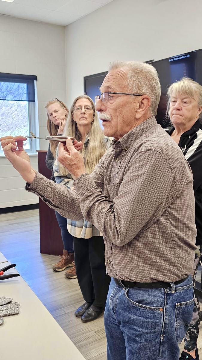Instructor demonstrates an apple tree grafting technique using a grafting knife and scion wood while participants watch during a fruit tree grafting workshop at the Clay County Center.