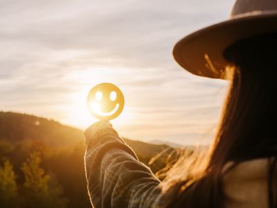 A person in a hat holds a round smiley-face disc toward the setting sun.