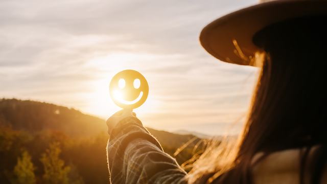 A person in a hat holds a round smiley-face disc toward the setting sun.