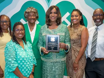 Six people pose in front of a large 4-H clover backdrop, with the woman at center holding the 2025 North Carolina 4-H Outstanding Citizen Alumni Award plaque.