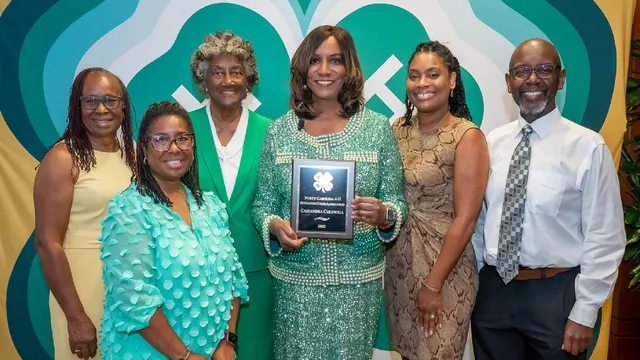 Six people pose in front of a large 4-H clover backdrop, with the woman at center holding the 2025 North Carolina 4-H Outstanding Citizen Alumni Award plaque.