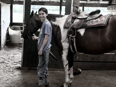 A smiling young rider in jeans and boots stands in a stable, holding the lead rope of a saddled brown-and-white pinto horse.
