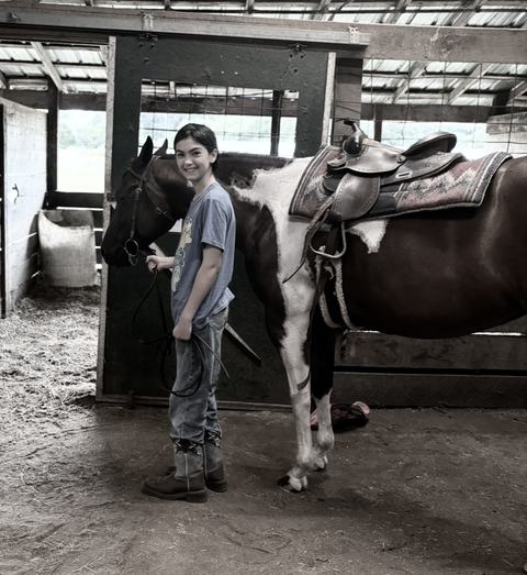 A smiling young rider in jeans and boots stands in a stable, holding the lead rope of a saddled brown-and-white pinto horse.